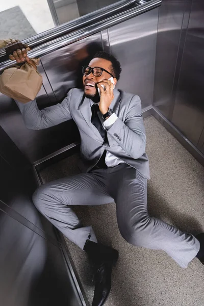 Crying african american businessman with paper bag talking on smartphone and suffering from panic attack in elevator — Stock Photo