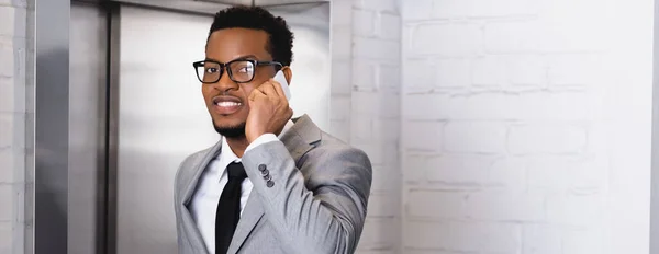 Plano panorámico de sonriente hombre de negocios afroamericano hablando en el teléfono inteligente mientras está de pie cerca del ascensor - foto de stock