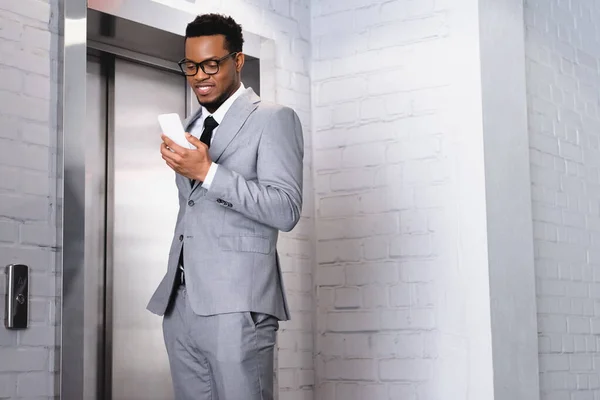 Улыбающийся африканский американский бизнесмен, держащий смартфон рядом с лифтом — стоковое фото