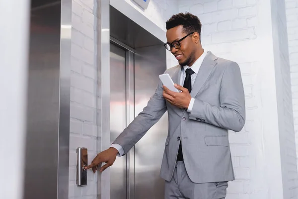 Uomo d'affari afro-americano positivo utilizzando lo smartphone mentre preme il pulsante dell'ascensore — Foto stock