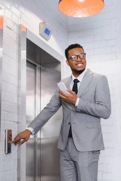 Sonriente hombre de negocios afroamericano sosteniendo teléfono inteligente y pulsando el botón del ascensor - foto de stock