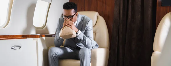 Panoramic shot of stressed african american businessman breathing with paper bag while having panic attack during flight on private plane — Stock Photo