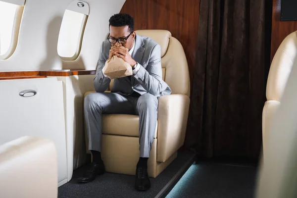 Homme d'affaires afro-américain nerveux respirant avec un sac en papier tout en ayant une attaque de panique pendant le vol en avion privé — Photo de stock