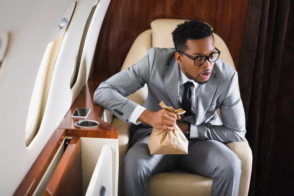 Worried african american businessman holding paper bag while having panic attack during flight on private plane — Stock Photo