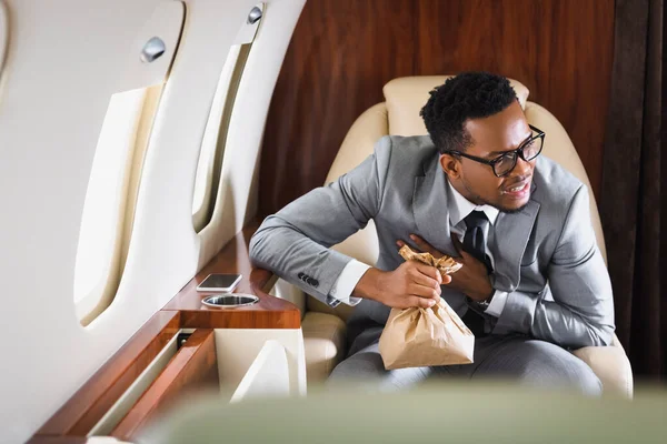 Nervous african american businessman holding paper bag while having panic attack during flight on private plane — Stock Photo