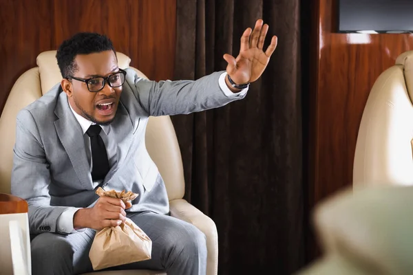 Homem de negócios afro-americano estressado com saco de papel tendo ataque de pânico durante o voo e mostrando gesto de parada — Fotografia de Stock