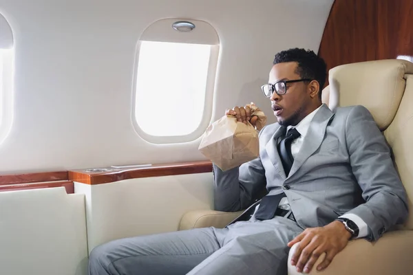 Preocupado hombre de negocios afroamericano respirando con bolsa de papel mientras tiene ataque de pánico durante el vuelo en avión privado - foto de stock