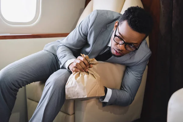 Nervous african american businessman holding paper bag while having panic attack during flight on plane — Stock Photo