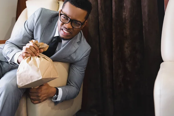 Estresado empresario afroamericano sosteniendo bolsa de papel mientras tiene ataque de pánico durante el vuelo en avión - foto de stock