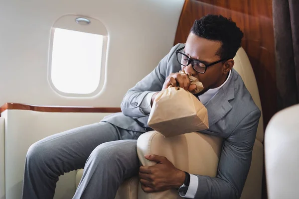 Homme d'affaires afro-américain nerveux respirant avec un sac en papier tout en ayant une attaque de panique pendant le vol en avion privé — Photo de stock