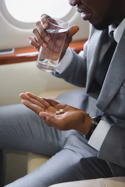 Vista recortada del hombre de negocios afroamericano nervioso tomando pastillas mientras tiene ataque de pánico durante el vuelo en avión privado - foto de stock