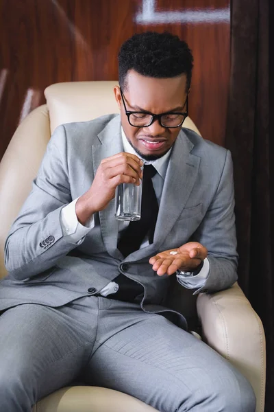 Hombre de negocios afroamericano nervioso tomando pastillas mientras tiene náuseas o ataque de pánico durante el vuelo en avión privado - foto de stock