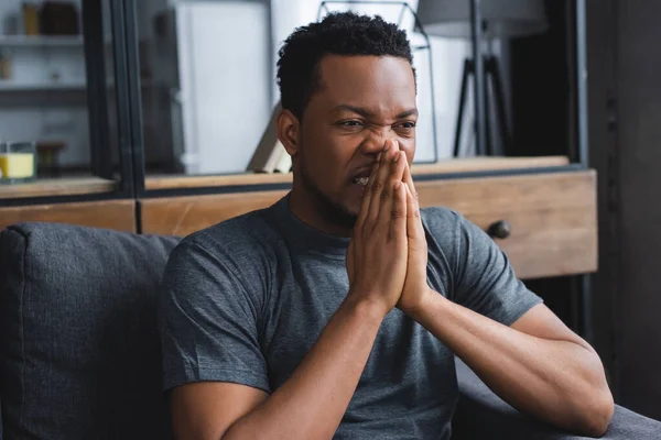 Homme afro-américain nerveux assis avec les mains ensemble sur le canapé à la maison — Photo de stock