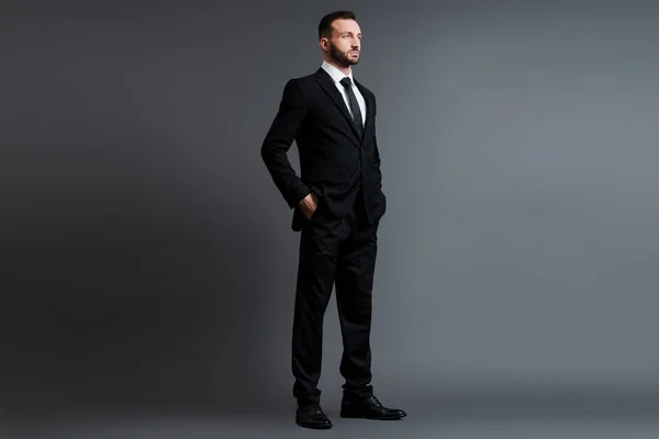 Homem de negócios bonito em terno de pé com as mãos nos bolsos em cinza — Fotografia de Stock