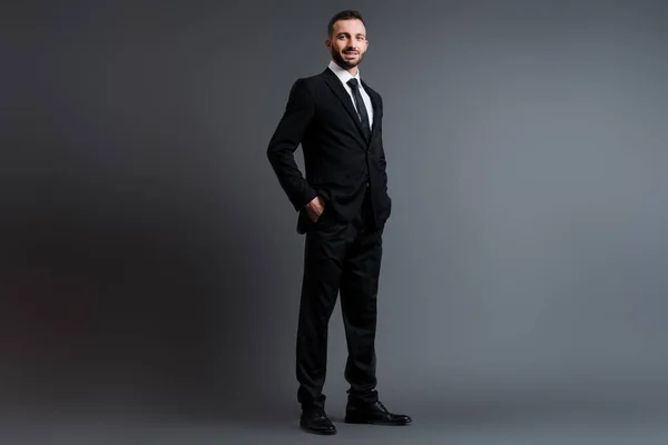 Glücklicher Geschäftsmann im Anzug mit Händen in Taschen auf grau — Stockfoto
