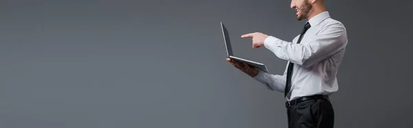 Plano panorámico de hombre de negocios en traje apuntando con el dedo a la computadora portátil aislado en gris - foto de stock