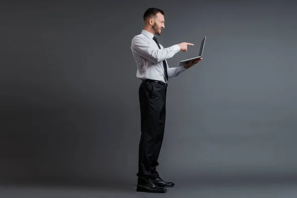 Profile of angry businessman in suit pointing with finger at laptop on grey — Stock Photo