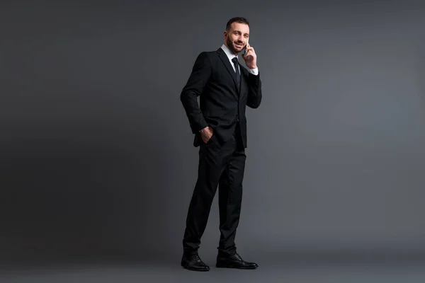 Alegre hombre de negocios en traje hablando en el teléfono inteligente, mientras que de pie con la mano en el bolsillo en gris - foto de stock