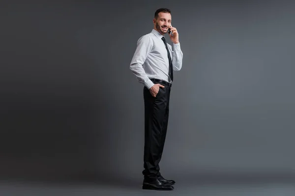 Усміхнений бізнесмен у костюмі говорить на смартфоні, стоячи з рукою в кишені на сірому — стокове фото