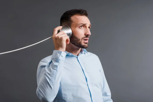 Bärtiger Mann mit Blechdose am Ohr beim Zuhören isoliert auf grau — Stockfoto