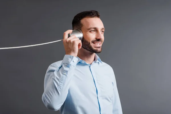 Hombre alegre sosteniendo lata puede cerca de la oreja mientras escucha aislado en gris - foto de stock