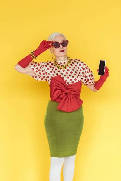 Stylish senior woman in sunglasses holding smartphone on yellow background — Stock Photo
