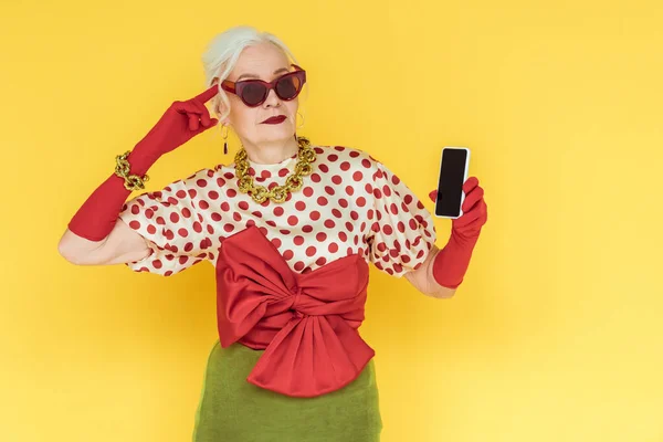 Fashionable senior woman pointing with finger on head and holding smartphone isolated on yellow — Stock Photo