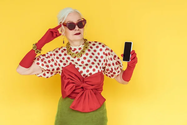 Fashionable senior woman pointing with finger on head and holding smartphone isolated on yellow — Stock Photo