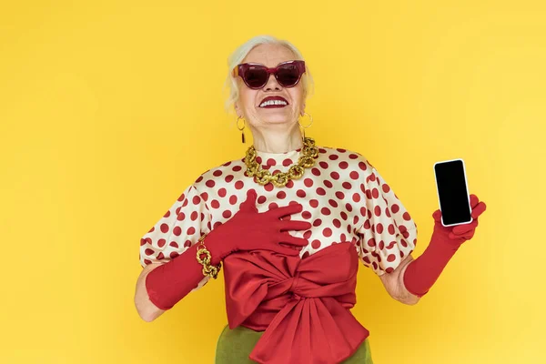 Модна старша жінка в сонцезахисних окулярах посміхається, тримаючи смартфон ізольовано на жовтому — стокове фото