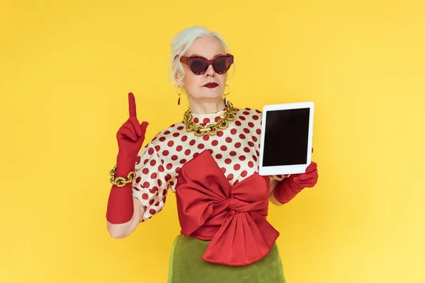 Stylish senior woman holding digital tablet and pointing with finger isolated on yellow — Stock Photo