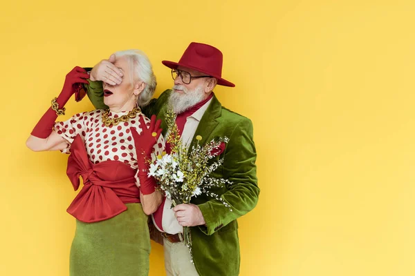 Un homme âgé élégant couvrant les yeux d'une femme et tenant un bouquet de fleurs sauvages sur fond jaune — Photo de stock