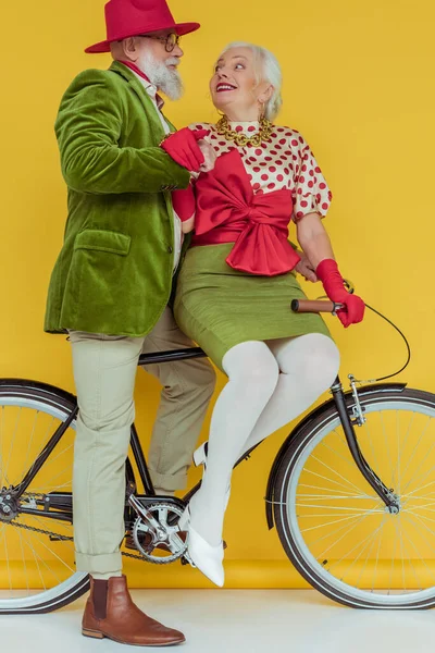 Pareja mayor de moda sonriéndose mutuamente en bicicleta sobre una superficie blanca sobre un fondo amarillo - foto de stock