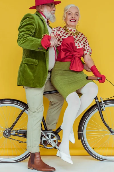 Mujer mayor sonriendo a la cámara cerca de hombre guapo en bicicleta en la superficie blanca sobre fondo amarillo - foto de stock