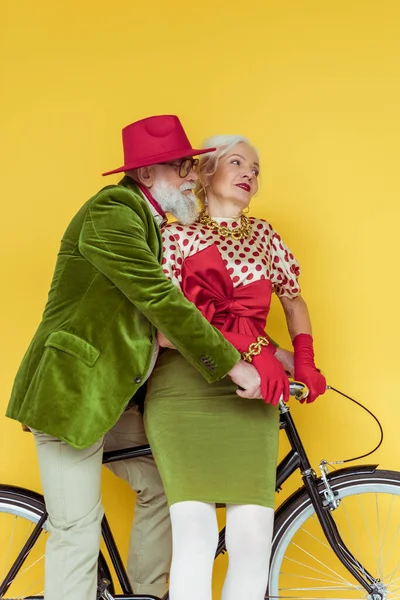 Pareja de ancianos de moda mirando hacia otro lado en bicicleta sobre fondo amarillo - foto de stock