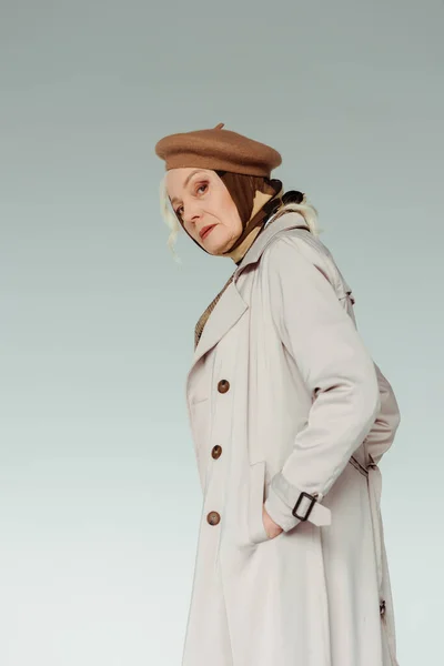 Side view of stylish senior woman looking at camera while holding hand in pocket of coat isolated on grey — Photo de stock