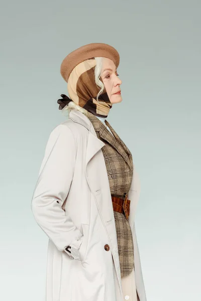 Side view of elegant elderly woman in coat and beret looking away isolated on grey — Stock Photo