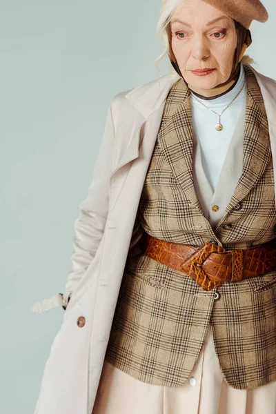 Femme âgée à la mode en manteau de tranchée regardant loin isolée sur grise — Photo de stock