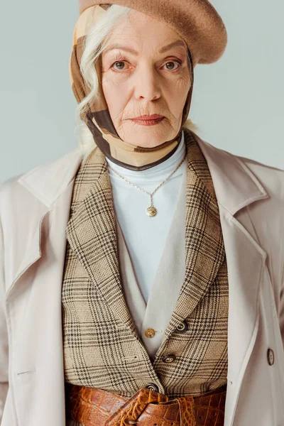 Une femme âgée élégante en béret et en manteau regarde une caméra isolée sur le gris — Photo de stock