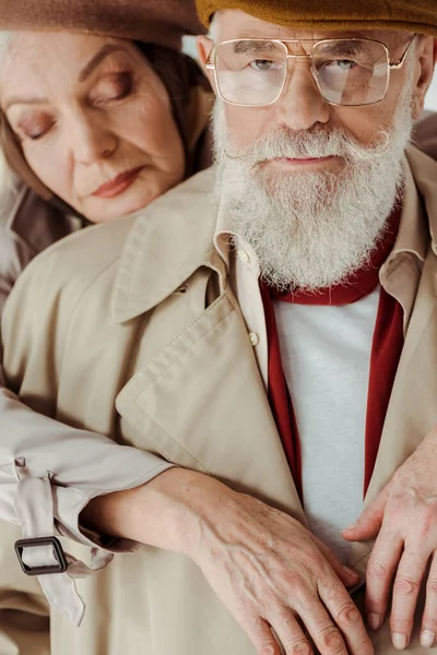 Selektiver Fokus eines eleganten älteren Mannes im Trenchcoat, der in die Kamera blickt, in der Nähe einer schönen Frau isoliert auf grau — Stockfoto