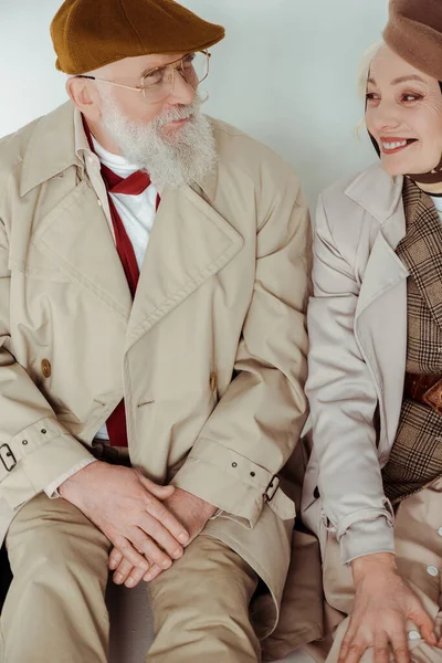 Stylish senior couple smiling at each other while sitting on white surface on grey background — Stock Photo