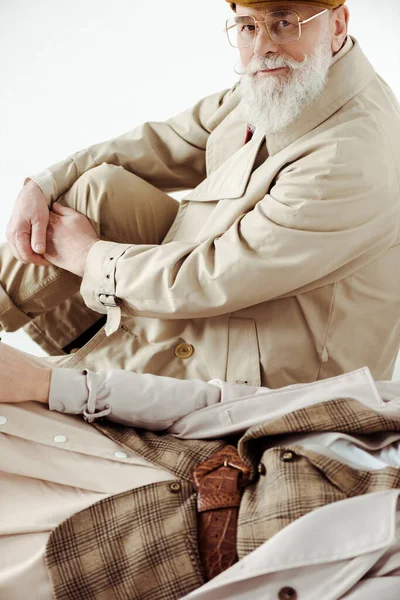 Vista lateral do homem sênior bonito em casaco de trincheira olhando para a câmera perto da mulher isolada no branco — Fotografia de Stock