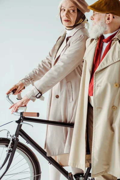 Vista lateral de la mujer de edad avanzada con estilo con la bicicleta mirando hombre elegante en gabardina aislado en blanco - foto de stock