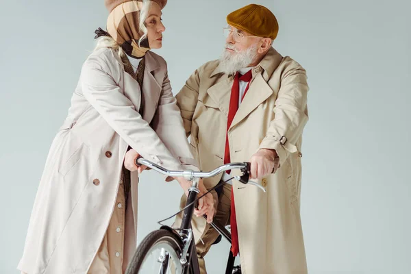 Elegante pareja de ancianos mirándose cerca de la bicicleta aislada en gris - foto de stock