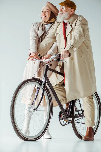 Volle Länge einer eleganten Seniorin, die in der Nähe eines gutaussehenden Mannes auf einem Fahrrad auf weißem Hintergrund weglächelt — Stockfoto