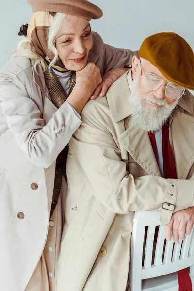 Sorridente donna anziana che abbraccia l'uomo alla moda sulla sedia isolata sul grigio — Foto stock