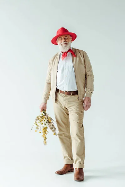 Pleine longueur d'un homme âgé élégant portant un chapeau rouge souriant à la caméra et tenant des fleurs sauvages sur fond blanc — Photo de stock