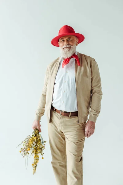 Hombre anciano de moda en sombrero rojo sosteniendo flores silvestres y sonriendo a la cámara aislada en blanco - foto de stock