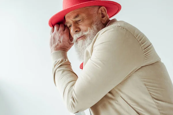 Vue latérale d'un homme âgé élégant portant un chapeau rouge, regardant ailleurs, assis sur une chaise, isolé sur un — Photo de stock