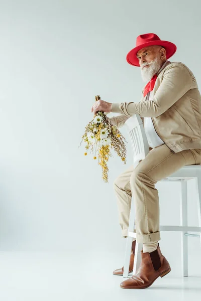 Повна довжина стильного літнього чоловіка в червоному капелюсі тримає букет з польових квітів, сидячи на стільці на білому тлі — стокове фото