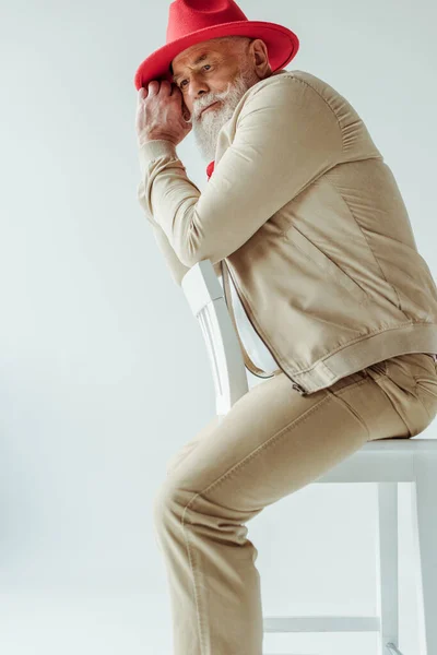 Side view of stylish senior man in red hat sitting on chair isolated on white — Stock Photo